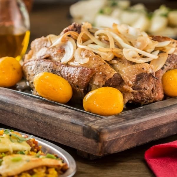 Variedade de petiscos e destaque à porção de carne assada com batatas, drinques e cervejas, em espaço animado.